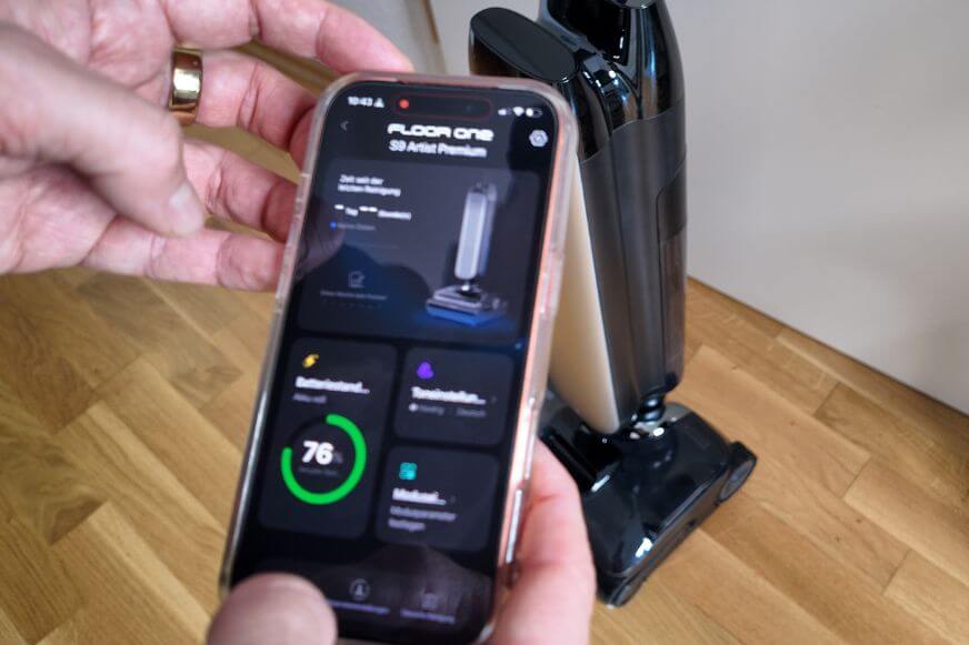 Tineco-App mit Akkustand und weiteren Infos, das Smartphone wird in der Hand gehalten und im Hintergrundsteht der Tineco Floor One S9 Artist.